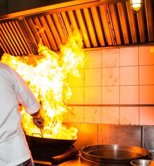 chef cooking with high open flames under commercial kitchen exhaust hood risk of grease fire requiring nfpa 96 compliance cleaning and fire suppression system maintenance