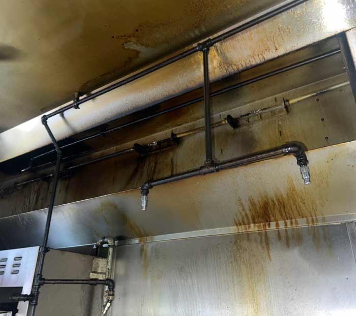 Grease-laden fire suppression pipes and stainless steel backsplash in a commercial kitchen requiring immediate hood cleaning service.