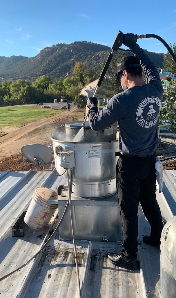 Technician pressure washing a rooftop upblast exhaust fan on a golf course clubhouse roof with mountains in the background