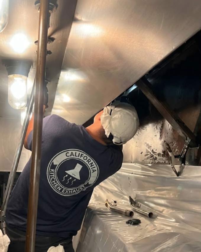 Technician scraping grease inside a restaurant exhaust hood with plastic sheeting protecting the cook line below